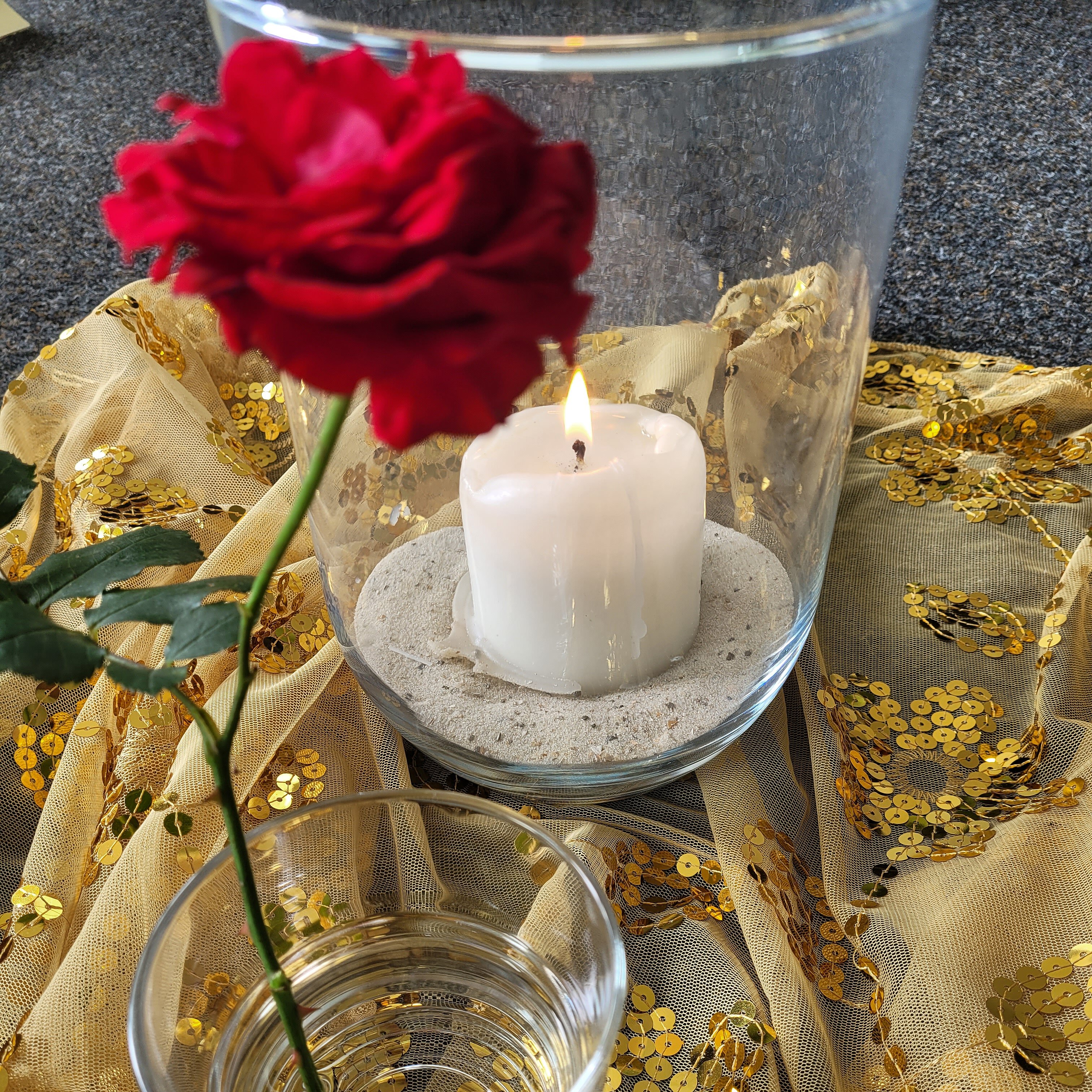 Zu sehen sind ein kleines Glas mit einer Rose und ein größeres Gefäß mit einer brennenden Kerze, die gemeinsam auf einem goldenen Tuch stehen.