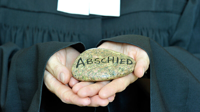 Ein Pastor hält einen Stein mit der Aufschrift "Abschied" in den Händen.