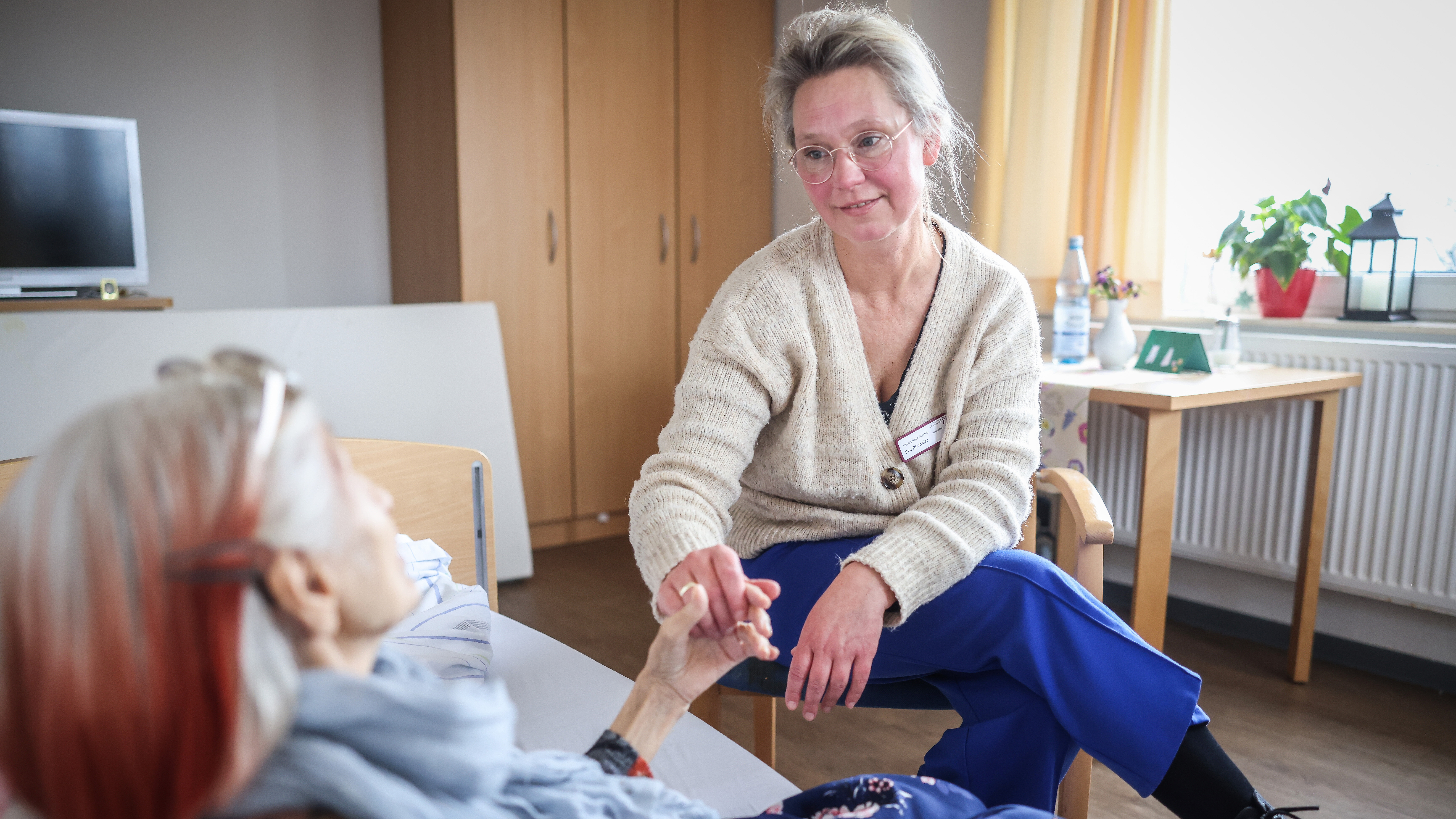 Mitarbeiterin sitzt auf einem Stuhl und hält die Hand einer älteren Frau, welche auf Ihrem Bett sitzt.