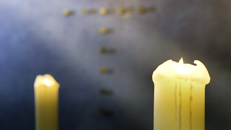 Zwei leuchtende Kerzen im Vordergrund, ein Kreuz im Hintergrund.
