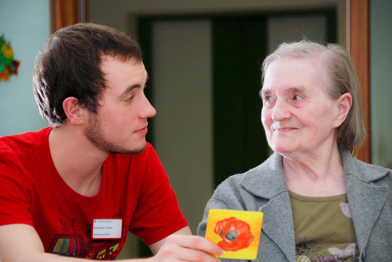 Ein Mitarbeiter sitzt zusammen mit einer älteren Frau an einem Tisch und spielt ein Spiel und hält dabei eine kleine Karte mit einer abgebildeten Mohnblume in der Hand.